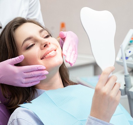 Woman in dental chair smiling at reflection with dentist’s gloved hands touching her cheeks