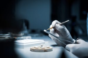 A few dental crowns placed on a tray while gloved hands hold one in a pair of forceps and are brushing it with a small instrument